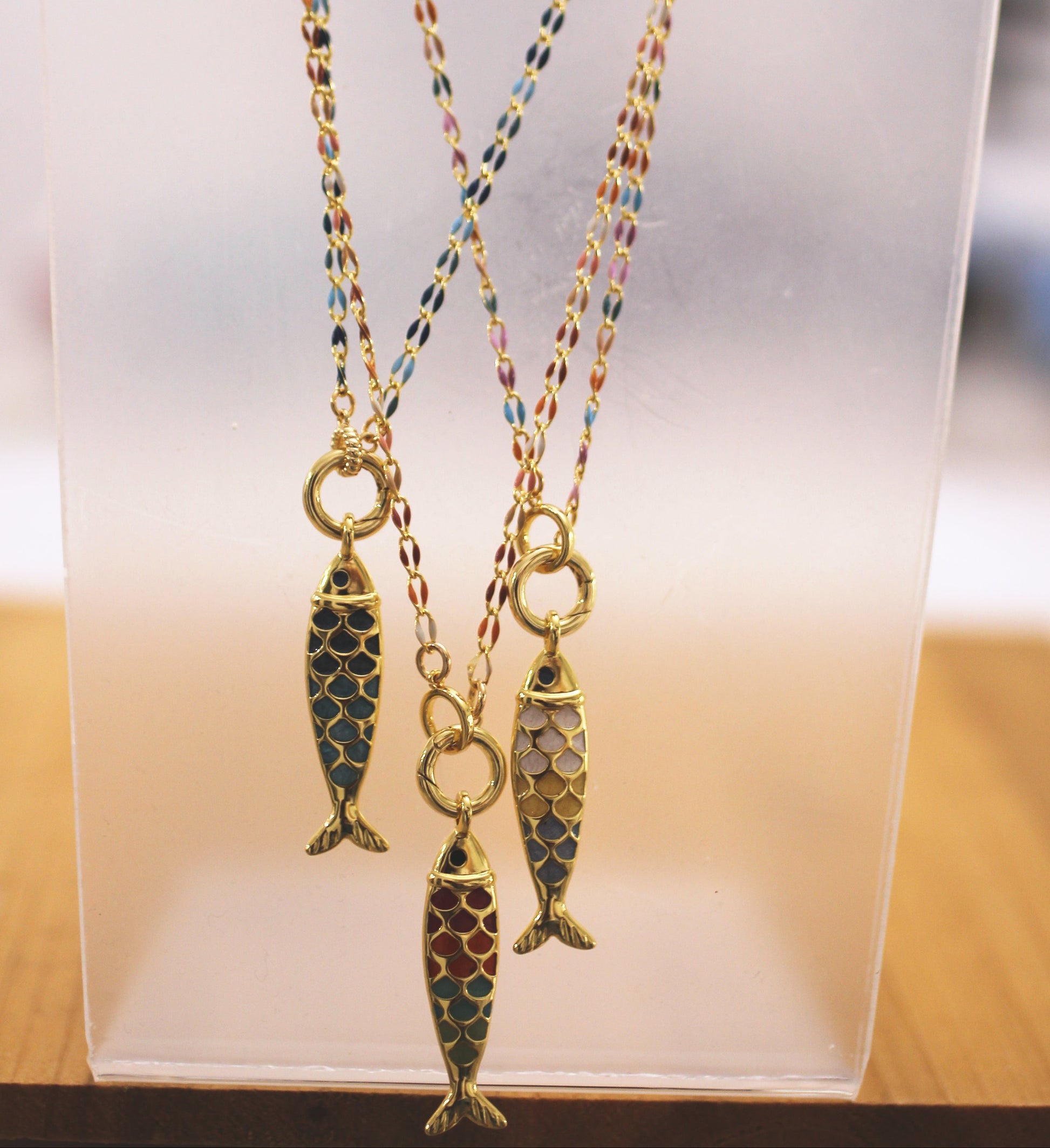 Three gold-tone fish pendant necklaces with colorful enamel scales, displayed on a clear acrylic stand.